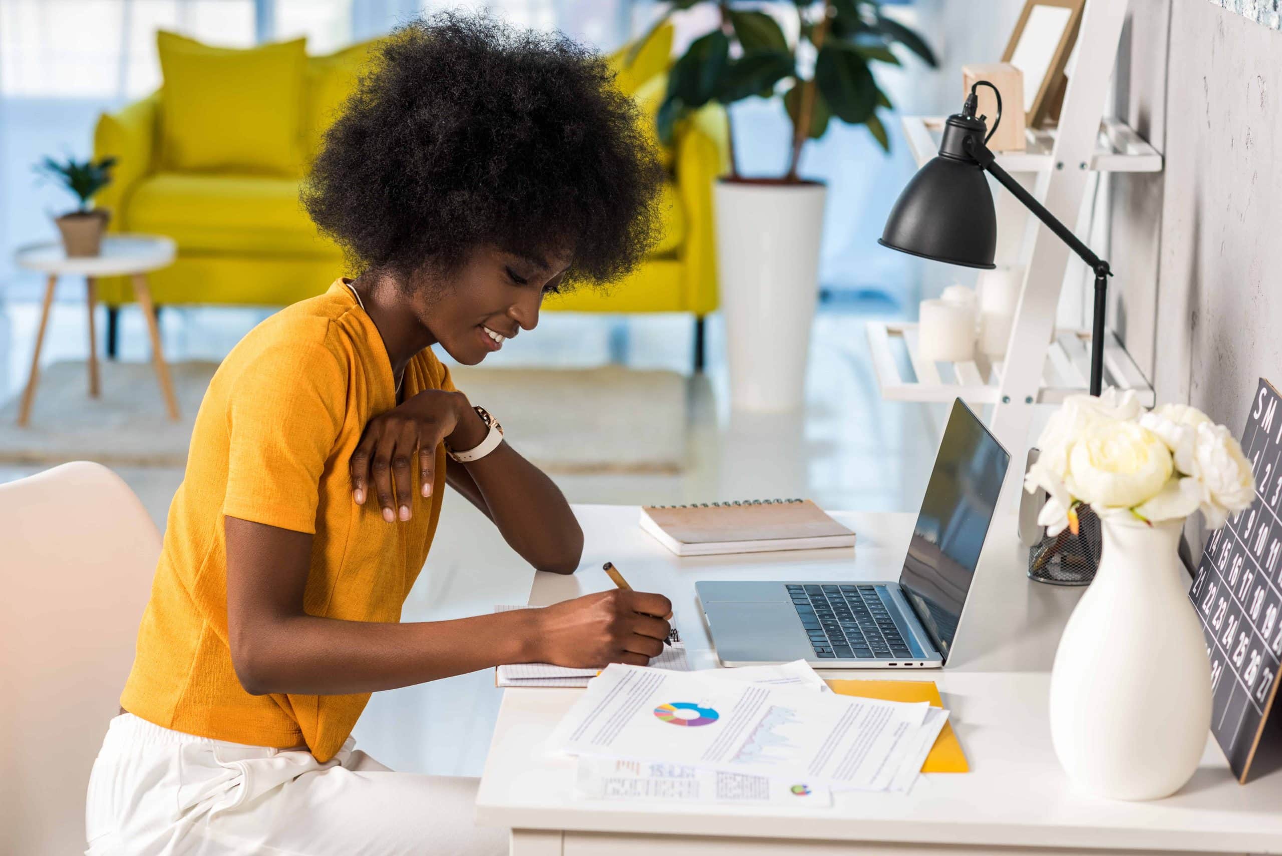 side view of a smiling freelancing woman. She is wearing a bright yellow shirt, and working at a desk with a laptop and papers.